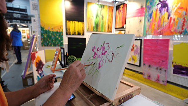 An artist paints pink flowers at a table with colorful abstract art displayed behind them.